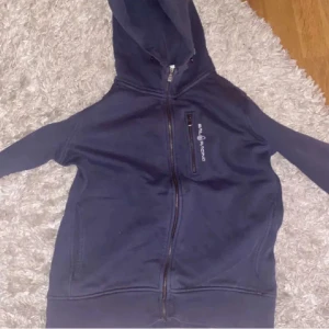 Mörkblå hoodie från Peak Performance - Mörkblå hoodie med dragkedja från Peak Performance. Tröjan har huva, fickor framtill och en diskret logga på bröstet. Perfekt för en avslappnad stil.