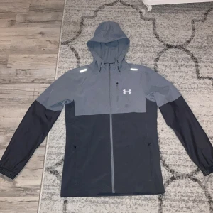 Grå och mörkblå vindjacka från Under Armour - Säljer en snygg tvåfärgad vindjacka från Under Armour i ljus och mörk grått. Jackan har huva, dragkedja framtill och två sidofickor. Perfekt för träning eller utomhusaktiviteter. Under Armour-logga på bröstet. Inga defekter, helt ny