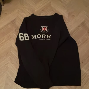 Svart långärmad tröja från Morris - Svart långärmad tröja från Morris med tryck på bröstet och siffran 68 på ärmen. Tröjan har rund halsringning och ett brittiskt flaggtryck framtill. 