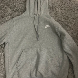 Grå hoodie från Nike - Säljer en klassisk grå hoodie från Nike med vit broderad logga på bröstet. Tröjan har huva med dragsko och en stor magficka framtill. Used men in bra skick.🤍