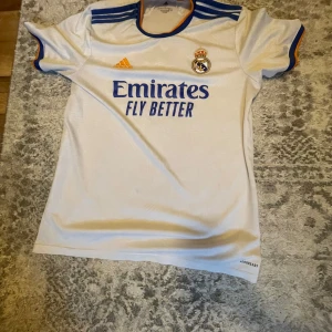 Real Madrid hemmatröja 2021/22 från Adidas - Vit Real Madrid fotbollströja från Adidas med blå och orange detaljer. Klassisk design med klubbmärke och sponsortryck på bröstet samt blå ränder på axlarna. Rund hals med orange kant. Perfekt för fans! Äkta från Real madrid shop