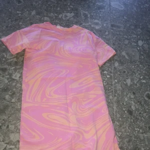 Rosa och orange mönstrad klänning från Lindex - Cool klänning från Lindex i rosa och orange med swirl-mönster och texten 'LET'S SMELL THE FLOWERS' på ryggen. Klänningen har korta ärmar och är perfekt för dig som gillar färg och statement-prints.