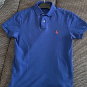 Blå pikétröja från Polo Ralph Lauren - Säljer en blå pikétröja från Polo Ralph Lauren med orange broderad logga på bröstet. Tröjan har klassisk krage, två knappar och är i slim fit-modell. Perfekt för en stilren och sportig look.