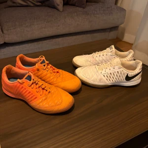 Nike fotbollsskor i orange och vitt - Två par Nike fotbollsskor, ett par i stark orange och ett par i vitt med svart Nike-logga. Skorna har snörning och är tillverkade i syntetmaterial med rund tå och platt sula. Perfekta för dig som gillar att spela fotboll och vill sticka ut på planen.