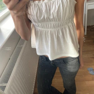 Vit linnetopp med smock och volang - Säljer en söt vit linnetopp med smockdetaljer och volangkant vid axelbanden. Toppen har en luftig och lätt passform, perfekt för varma dagar. Materialet känns mjukt och bekvämt mot huden. Enkel att matcha med både jeans och kjol.💕🌸
