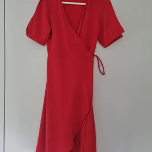 Röd omlottklänning från Pieces - Säljer en röd omlottklänning från Pieces i storlek XS. Klänningen har korta puffärmar, knyt i midjan och en snygg v-ringning. Perfekt för dig som gillar en feminin och stilren look. Finns liten fläck som kan anas vid omlottet fram