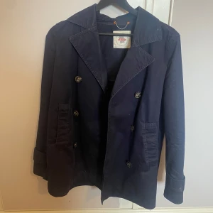 Marinblå vår/sommar jacka med knappar - Säljer en klassisk marinblå trenchcoat med dubbelknäppning. Perfekt för dig som gillar tidlös stil och vill ha en jacka som funkar till många tillfällen. Storlek S