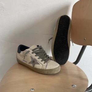 Vita sneakers med grå stjärndetalj - Säljer ett par vita sneakers från Golden Goose med grå snörning och grå stjärna på sidan. Skorna har blå detalj vid hälen och beige sula. Perfekta för en avslappnad och trendig stil. De är till 100 procent äkta och jag har box till dem. Har använt dem 4 gånger. 