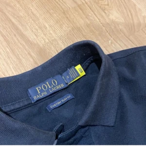 Svart pikétröja från Polo Ralph Lauren - Klassisk svart pikétröja från Polo Ralph Lauren med röd broderad logga på bröstet. Tröjan har korta ärmar, krage och knäppning framtill. Modellen är custom slim fit och passar perfekt till en stilren look.
