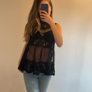 Svart spetsblus med volang - Säljer en svart, ärmlös blus i transparent spets med blommigt mönster och volang nertill. Blusen har en lös passform och är perfekt för lager-på-lager-stil. Supersnygg att styla med jeans eller kjol. Köpte 1 år sedan.