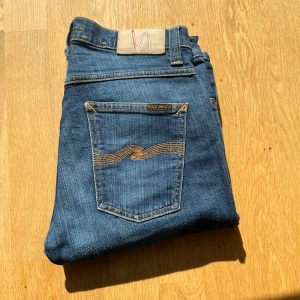 Riktigt fräscha blåa ”Grim Tim” Nudie Jeans - Säljer ett par klassiska blå jeans från Nudie Jeans med orange sömmar och snygg broderad detalj på bakfickorna. Jeansen har normal passform och raka ben. Perfekta till vardags för en stilren look. Storlek : W:30 L:32 Kom gärna med offer🙌