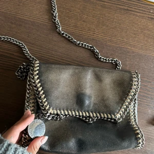 Vintage Stella McCartney - Cool svart axelväska från Stella McCartney med silverfärgad kedja som axelrem och runt kanterna. Väskan har lock med magnetknäppning och insida i ljusrosa tyg. Perfekt för dig som vill ha en snygg accessoar med statement-detaljer.