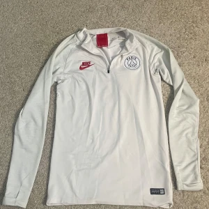 Vit långärmad PSG fotbollströja från Nike - Säljer en vit långärmad Paris Saint-Germain fotbollströja från Nike. Tröjan har röd Nike-logga på bröstet och PSG-emblem på andra sidan. Dragkedja vid halsen och autentisk football-märke nertill. Perfekt för träning eller match.