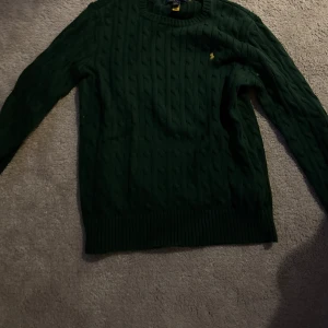 Mörkgrön stickad tröja från Polo Ralph Lauren - Mörkgrön stickad tröja från Polo Ralph Lauren Säljer en mörkgrön stickad tröja från Polo Ralph Lauren med klassiskt kabelstickat mönster och rund halsringning. Tröjan har långa ärmar och en liten gul broderad logga på bröstet. Perfekt för kyligare dagar.