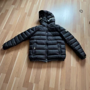 Svart dunjacka från Moncler med huva - Säljer en svart dunjacka från Moncler med huva och dragkedja. Jackan har långa ärmar, två framfickor med knappar och klassisk Moncler-logga på ärmen. Snygga vita detaljer på huvan ger en unik look. Även perfekt för kyliga dagar på kvällarna under vår eller sommar.