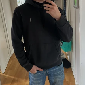 Svart hoodie från Polo Ralph Lauren - Säljer en svart hoodie från Polo Ralph Lauren med klassisk logga på bröstet. Tröjan har huva med dragsko och en stor magficka framtill. Perfekt för en avslappnad och stilren look.
