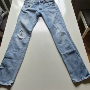 Ljusblå jeansbyxor från Replay - Säljer ett par ljusblå jeansbyxor från Replay med klassisk femficksdesign och diskreta slitningar på vänster lår. Byxorna har raka ben och normal passform, samt detaljerad söm på bakfickan. Perfekta för en avslappnad stil.