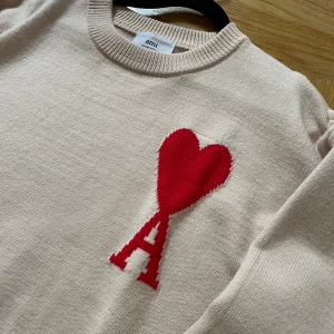 Beige stickad tröja med rött hjärta från Ami - Säljer en beige stickad tröja från Ami med ett stort rött hjärta och bokstaven A på bröstet. Tröjan har rund halsringning, långa ärmar och ribbade muddar vid ärmslut och nederkant. Perfekt för dig som gillar stilrena detaljer.