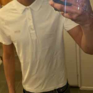 Vit piké t-shirt från Hugo Boss - Säljer en vit piké t-shirt från Hugo Boss med guldigt BOSS-tryck på bröstet. Slim fit-modell med klassisk krage och knappar framtill. Perfekt för en clean och stilren look.