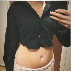 Svart croppad skjorta - Säljer en svart croppad skjorta med klassisk krage och långa ärmar. Skjortan har knappar framtill och är perfekt för en trendig och avslappnad stil.  skit snygg till några kostymbyxor eller jeans😘