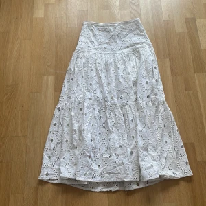 Vit broderad långkjol från Pink Woman - Superfin vit långkjol från Pink Woman med broderade blommor och hålmönster. Kjolen har volanger och dragkedja i sidan. Perfekt för sommarens alla tillfällen!🤍