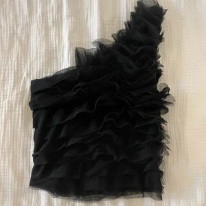 Svart one shoulder topp med volanger - Säljer en svart one shoulder topp med flera lager volanger i tyll. Toppen har en asymmetrisk design och är perfekt för dig som vill ha något unikt.
