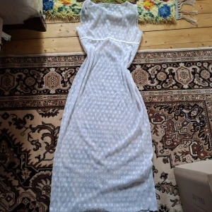 Vit långklänning med blommigt mönster - Säljer en vit långklänning med diskret blommigt mönster. Klänningen är ärmlös och har en markerad midja med en lätt figurnära passform. Perfekt för sommarens alla tillfällen.