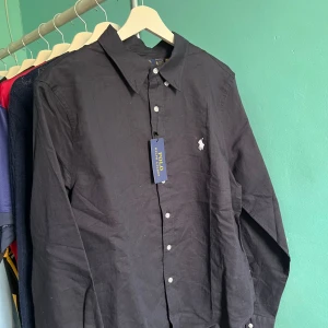 Polo Ralph Lauren Linneskjorta - Klassisk marinblå skjorta från Polo Ralph Lauren med vit broderad logga på bröstet. Skjortan har knappar framtill och är långärmad. Perfekt för en stilren och tidlös look.