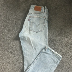 Levi's 501 ljusblå jeans - Säljer ett par klassiska Levi's 501 jeans i ljusblå tvätt. Byxorna har raka ben, knäppning med knappar och fem fickor. Perfekta för en avslappnad och tidlös stil. Levi's Premium-märke på insidan och läderpatch bak. Har ett litet märke i tyget, se sista bilden. 