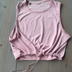 GYMSHARK  BREEZE TOP (M) - Säljer en ljusrosa croppad topp från Gymshark. Toppen har rund halsringning, ärmlös design och dragsko i nederkanten för justerbar passform. Perfekt för träning eller en sportig look.