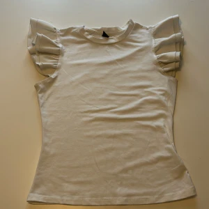 Vit topp med volangärmar från SHEIN - Säljer en vit topp från SHEIN med korta volangärmar som ger en söt och trendig look. Toppen har rund halsringning och är tillverkad i ett mjukt material. Perfekt att styla till jeans eller kjol!