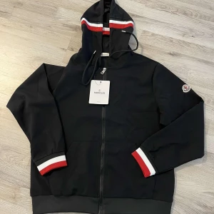 Svart hoodiejacka från Moncler med rödvita detaljer - Svart hoodiejacka från Moncler med dragkedja framtill. Huva och ärmslut har snygga rödvita ränder. Klassisk Moncler-logga på ärmen och ribbade muddar. Perfekt för en sportig och stilren look.