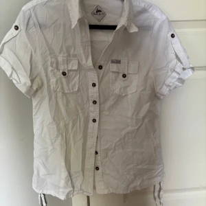 Vit kortärmad skjorta från Blanc du Nil - Säljer en vit skjorta från Blanc du Nil med korta ärmar, knappar framtill och två bröstfickor med lock. Skjortan har knappar på ärmarna för att kunna vika upp dem och dragsko nertill för justerbar passform.