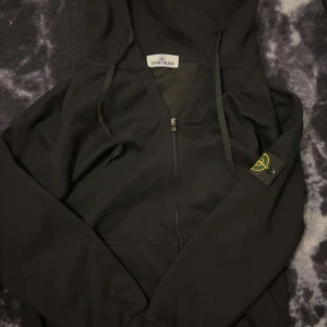 Svart hoodiejacka från Stone Island - Svart hoodiejacka från Stone Island med dragkedja framtill och klassisk logotyp på ärmen. Jackan har huva med dragsko och en stilren, sportig look. Perfekt för dig som gillar streetwear och vill ha något bekvämt och snyggt. Pris kan diskuteras vid snabb affär