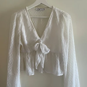 Vit blus med knyt framtill från NA-KD - Säljer en söt vit blus från NA-KD med knyt framtill och v-ringning. Blusen har långärmade, luftiga ärmar och ett diskret prickigt mönster i tyget. Perfekt att styla med jeans eller kjol för en trendig look. Storlek 36, men känns som en M. 