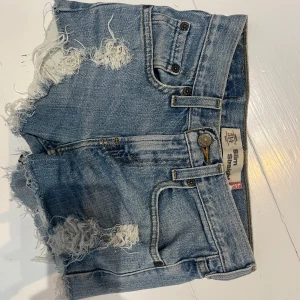 Blå jeansshorts från Levi's - Säljer ett par klassiska blå jeansshorts från Levi's med slitna detaljer och fransar. Shortsen har låg midja, fem fickor och råa kanter. Perfekta för sommaren och ger en cool vintagekänsla.