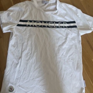 Vit t-shirt från Moncler med logga - Vit t-shirt från Moncler med svart logga och två svarta ränder över bröstet. Klassisk rund halsringning och korta ärmar. Liten Moncler-patch nertill på framsidan. Perfekt för en stilren och enkel look. Användning vid fåtal sekvenser men texten är dock lite förstörd pga tvättning men den är fortfarande lika fräsch som från när jag köpte den 