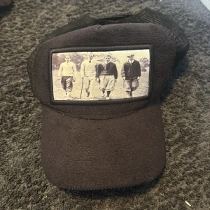 Svart keps från Emilio Pendenza med vintagefoto - Svart keps med mesh-bak och justerbar snapback. Framtill finns ett vintageinspirerat foto av fyra personer. Märkt med Emilio Pendenza på insidan och baksidan. Perfekt för en avslappnad stil.