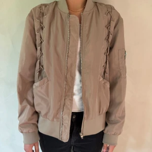 Beige bomberjacka med snörningsdetaljer - Säljer en beige bomberjacka med snygga snörningsdetaljer längs axlarna och ärmarna. Jackan har dragkedja framtill, ribbade muddar och fickor på sidorna samt en ärmficka med dragkedja. Perfekt för dig som vill ha en unik twist på klassikern.