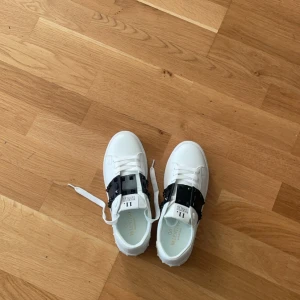 Valentino Garavani Open sneakers vit/svart - Pris kan diskuteras. Snygga Valentino Garavani Open sneakers i vitt skinn med svart band över vristen och klassiska nitar på sulan. Skorna har rund tå, platt sula och vita snören. Perfekta för dig som vill ha en lyxig och trendig look.