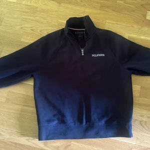 Mörkblå Tommy Hilfiger half zip tröja - Snygg mörkblå tröja från Tommy Hilfiger med broderad logga på bröstet. Modellen har half zip-dragkedja och hög krage med klassisk rödvit detalj på insidan. Perfekt för dig som gillar stilren och sportig look.