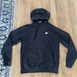 Svart Nike hoodie med broderad logo - Svart hoodie från Nike med klassisk broderad vit logga på bröstet. Tröjan har huva med snörning och en stor känguruficka framtill. Tillverkad i mjuk bomullsmix och har långa ärmar med muddar. Perfekt för en chill och sportig stil.