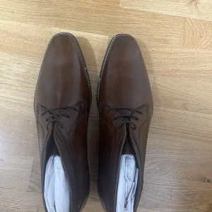 Bruna skinnkängor med snörning - Snygga bruna kängor i skinn med klassisk snörning och fyrkantig tå. Perfekta för dig som vill ha en stilren och tidlös look. Skorna har en låg klack och platt sula, vilket gör dem bekväma att bära hela dagen.