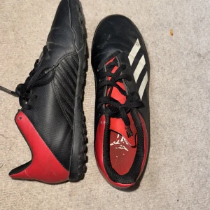 Svarta och röda adidas fotbollsskor - Säljer ett par adidas fotbollsskor i svart och rött med klassiska vita ränder på sidan. Skorna har snörning och en greppvänlig sula med små dobbar, perfekt för spel på konstgräs. Tillverkade i syntetmaterial och har en sportig look.