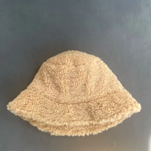 Beige teddy bucket hat - Supermjuk och fluffig bucket hat i beige teddy-material. Hatten har en klassisk form med bred brätte och är fodrad med vitt tyg för extra komfort. Perfekt accessoar för att lyfta din höst- eller vinterstil.
