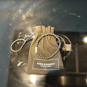Svart pärlarmband Steel & Barnett - Stilrent svart armband från Steel & Barnett med matta pärlor och en diskret metallbricka med logga. Armbandet levereras i en snygg svart tygpåse med dragsko, perfekt att ge bort eller ha själv. Alldrig använt 