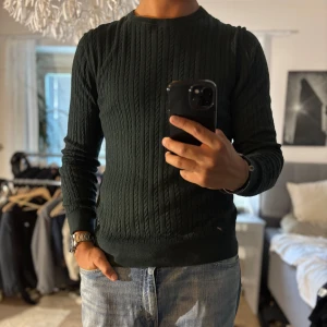 Mörkgrön stickad tröja Jack & Jones - Snygg mörkgrön stickad tröja från Jack & Jones Premium. Tröjan har ett klassiskt kabelstickat mönster, rund halsringning och långa ärmar. Perfekt för dig som gillar en stilren och enkel look med lite struktur. Passar till både jeans och chinos.