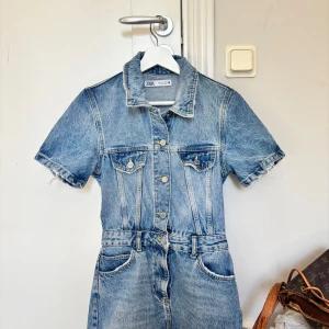 Jeansklänning med kort ärm från Zara - Snygg jeansklänning från Zara i klassisk blå denim. Modellen har korta ärmar med råa kanter, knappar framtill, bröstfickor och markerad midja. Perfekt för dig som gillar en cool och avslappnad stil.