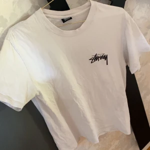 Vit Stüssy t-shirt med logga - Vit t-shirt från Stüssy med klassisk svart logga tryckt på bröstet. Enkel och clean design med rund hals och korta ärmar. Perfekt för dig som gillar streetwear och vill ha en stilren look.