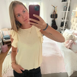Ljusgul blus med volangärmar - Supersöt ljusgul blus med volangdetaljer på axlarna och rund halsringning. Blusen är ärmlös och har en snygg öppning i ryggen med knapp. Perfekt för dig som gillar en feminin och luftig stil. Materialet känns lätt och flowy.